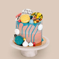 The Kids Works Cake by Jenna Rae Cakes features blue drip icing, macarons, meringues, candy balls, and a Happy Birthday sign, displayed on a white cake stand against a beige background.