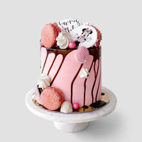 The Kids Works Cake by Jenna Rae Cakes is a pink cake with chocolate drip, topped with pink macarons, meringues, candy, and a white topper featuring a cartoon mouse and “Happy B,” displayed on a white stand against a plain backdrop.