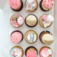 The Works Cupcakes by Jenna Rae Cakes: a box of 12 assorted cupcakes topped with macarons, gold accents, sprinkles, meringue, and pink flower-shaped icing. One cupcake features Happy Birthday in black script.