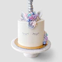 The Unicorn Cake by Jenna Rae Cakes features white frosting, closed eyes on the side, pastel flowers and ears on top, plus a silver unicorn horn, beautifully presented on a round marble cake stand.