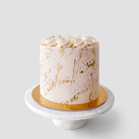 The Vanilla Cake by Jenna Rae Cakes features white frosting with gold splatters and cream swirls, presented on a gold board and white marble stand against a plain light background.