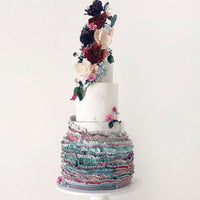 A three-tiered cake with textured, ruffled layers in pastel colors and decorated with cascading, realistic flowers in shades of pink, purple, and white, displayed on a white cake stand against a plain background.