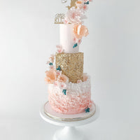 A three-tiered cake with pink, gold, and white layers, decorated with ruffled icing, pastel flowers, a gold cupid topper, and blue butterfly decorations, displayed on a white cake stand against a white background.