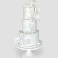 A three-tiered white cake decorated with delicate, white sugar flowers cascading down the side, displayed on a simple white cake stand against a plain light gray background.