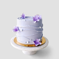 Discover Jenna Rae Cakes Wedding Cakes Under $200: a round, light purple cake adorned with wavy icing lines, white sugar pearls, and purple sugar flowers—beautifully displayed on a marble stand with a gold base.