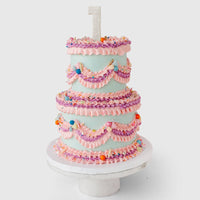 The Wedding Cakes Under $500 by Jenna Rae Cakes features three tiers, pink and purple icing swags, colorful sprinkles, a glittery number one candle topper, and comes displayed on a white cake stand.