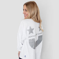A woman with long blonde hair smiles over her shoulder, wearing the Jenna Rae Cakes White and Silver Spirit Jersey with two stars on the back, paired with black shorts.