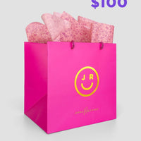 The $100 JRC Gift Bag by Jenna Rae Cakes features a bright pink bag with a yellow smiley face logo and branding, filled with pink tissue paper. $100 appears in bold purple text at the top right corner.