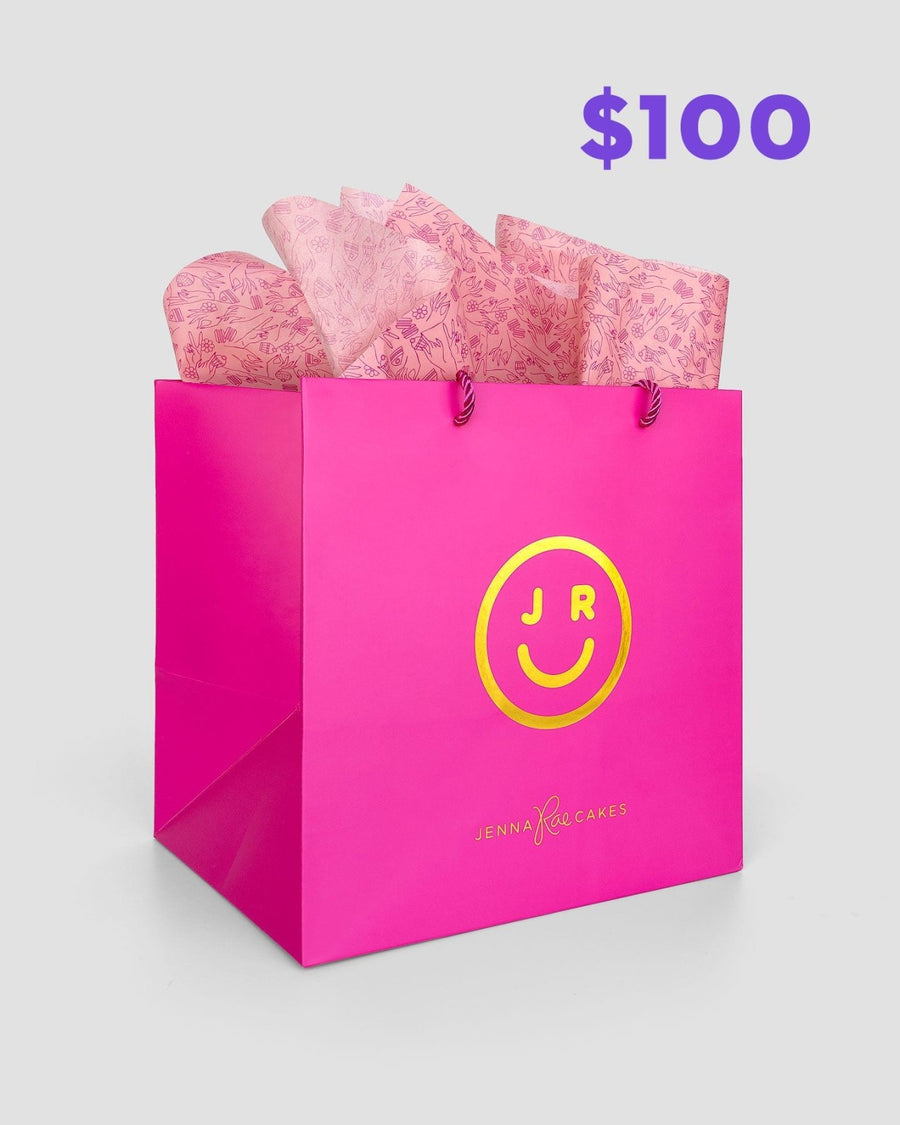 The $100 JRC Gift Bag by Jenna Rae Cakes features a bright pink bag with a yellow smiley face logo and branding, filled with pink tissue paper. $100 appears in bold purple text at the top right corner.
