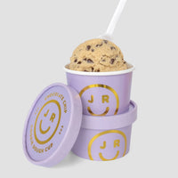 A $100 JRC Gift Bag from Jenna Rae Cakes in a purple cup with a white spoon, stacked on another matching lidded cup. Both cups show gold smiley face logos and text.