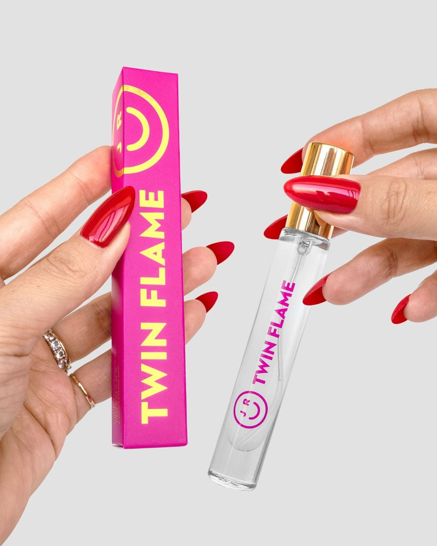 A person with red manicured nails holds a pink and yellow $165 JRC Gift Bag box by Jenna Rae Cakes in one hand and a clear bottle labeled $165 JRC Gift Bag in the other.