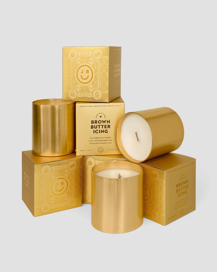 A stack of Jenna Rae Cakes $165 JRC Gift Bags with gold candle jars and matching gold boxes, labeled Brown Butter Icing, arranged neatly against a light gray background.