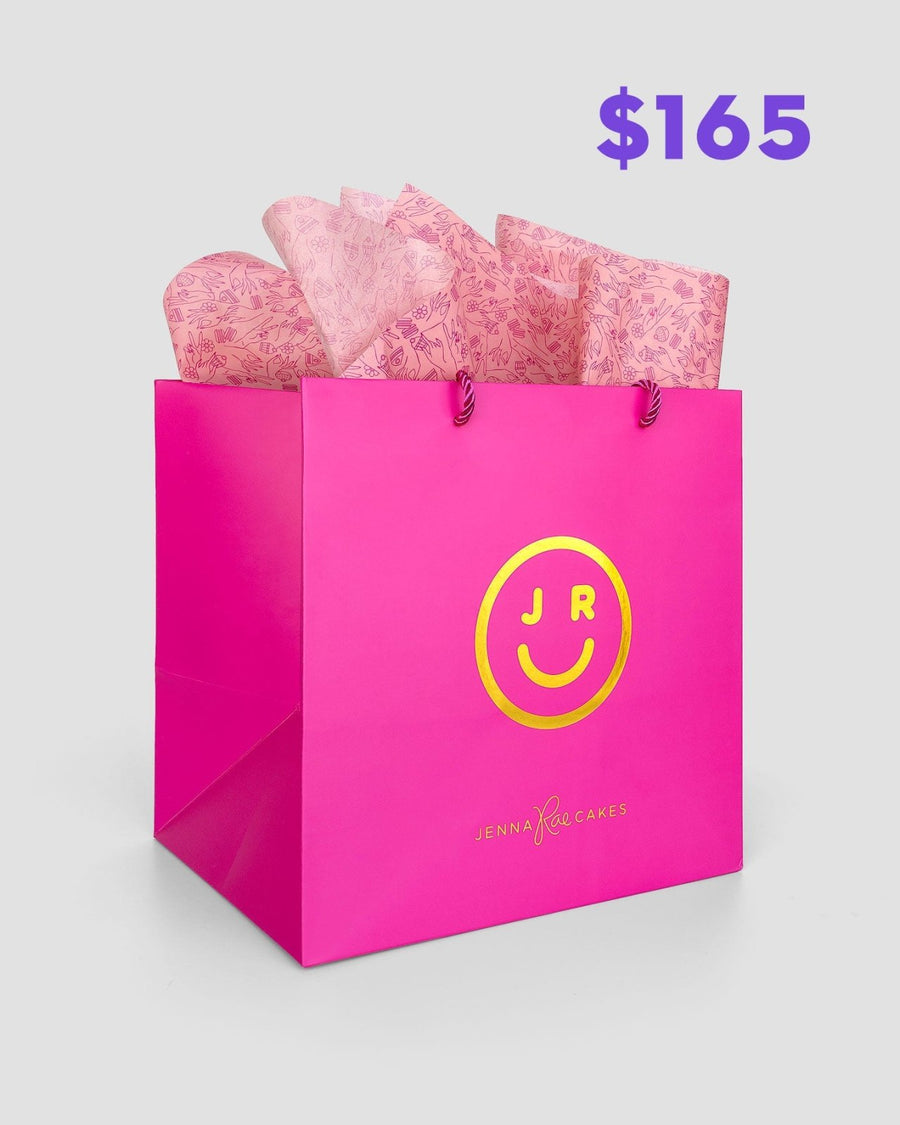 The $165 JRC Gift Bag by Jenna Rae Cakes features a bright pink bag with a yellow smiley face logo and is filled with pink patterned tissue paper. Jenna Rae Cakes appears on the bag, and purple $165 text is shown in the top right corner.