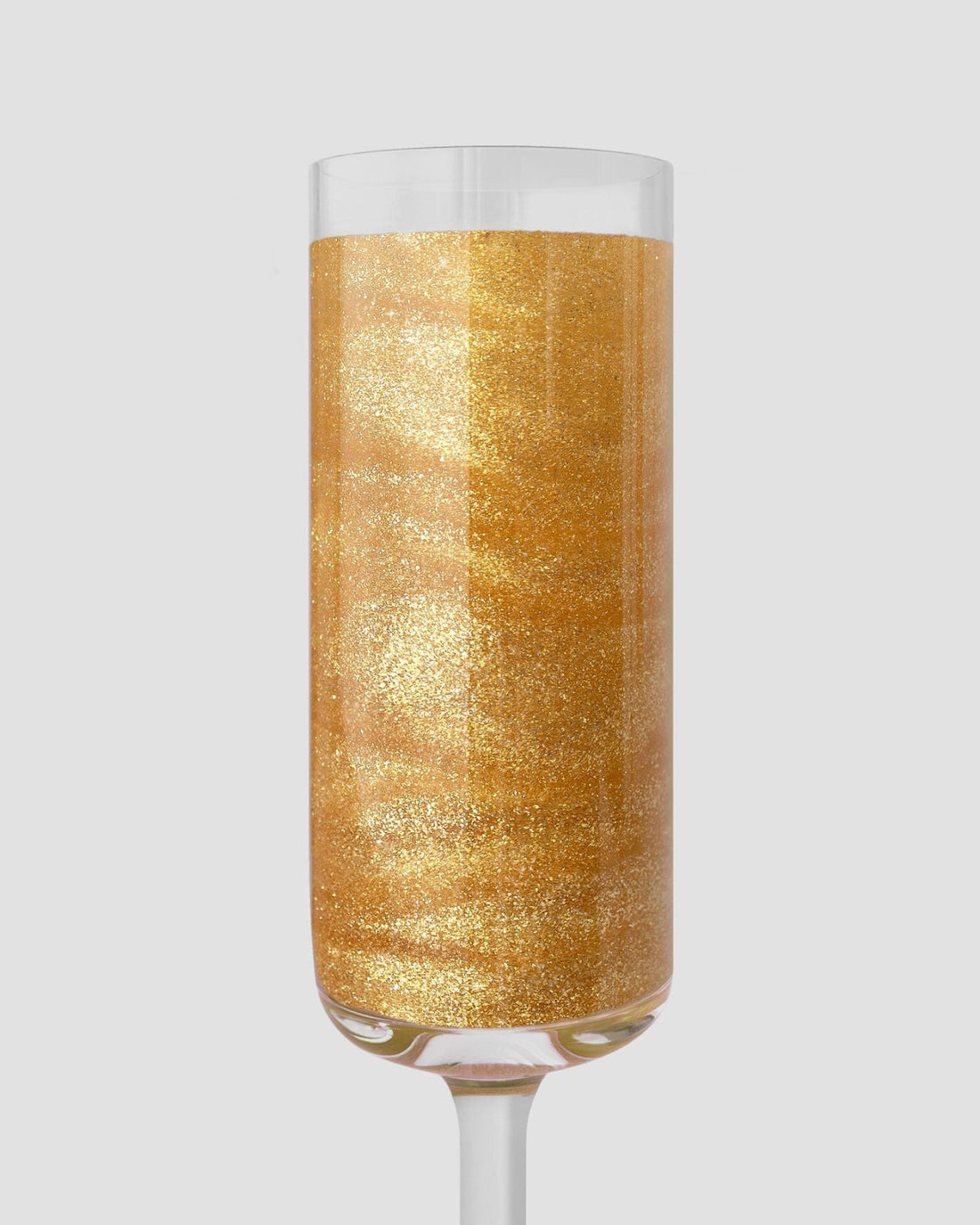 A clear champagne flute filled with golden FND TESTING - 24k Magic Edible Glitter by Jenna Rae Cakes shimmers against a plain light gray background.