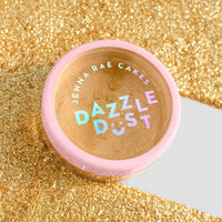 A small round container with a pink lid labeled Jenna Rae Cakes FND TESTING - 24k Magic Edible Glitter sits on sparkling gold glitter, featuring colorful, holographic letters on its label.