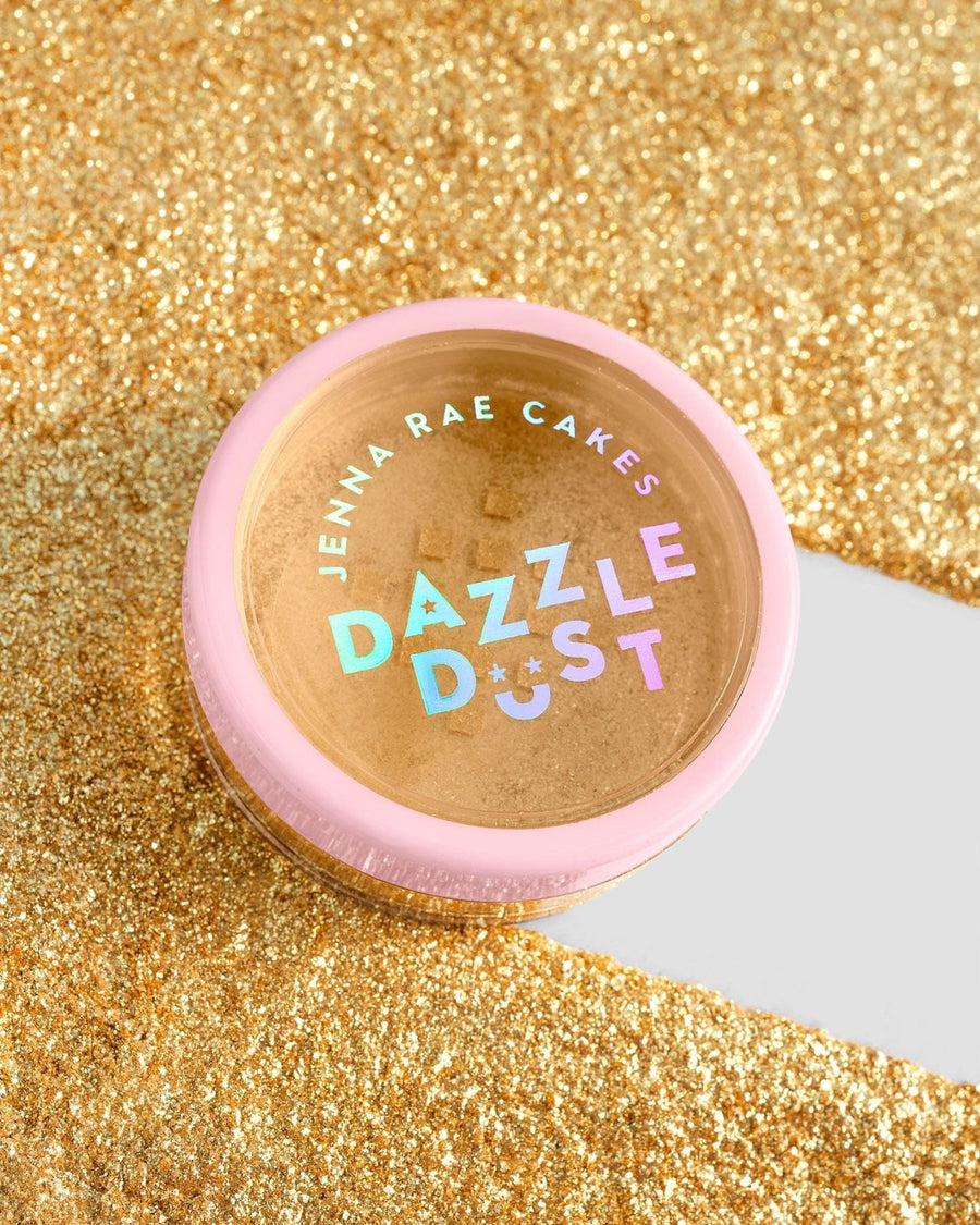 A small round container with a pink lid labeled Jenna Rae Cakes FND TESTING - 24k Magic Edible Glitter sits on sparkling gold glitter, featuring colorful, holographic letters on its label.