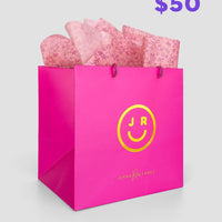 The $50 JRC Gift Bag by Jenna Rae Cakes features a bright pink bag with a yellow smiley face logo and brand text, filled with pink patterned tissue paper, set against a light grey background.