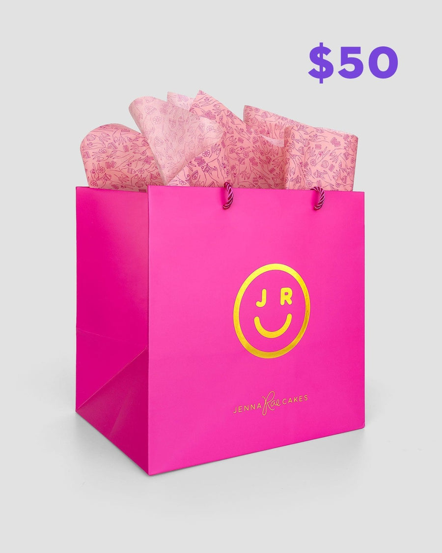 The $50 JRC Gift Bag by Jenna Rae Cakes features a bright pink bag with a yellow smiley face logo and brand text, filled with pink patterned tissue paper, set against a light grey background.