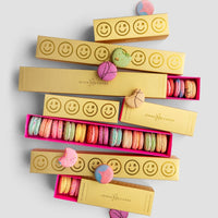 $75 JRC Gift Bags by Jenna Rae Cakes, featuring gold boxes with smiley faces and bright pink interiors, are filled with colorful macarons, some displayed outside the boxes on a white surface.
