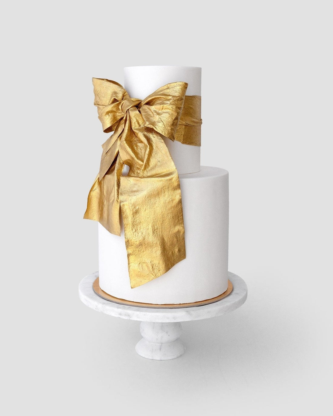 A two-tier white cake on a marble stand features a large bow on the top tier, shimmering with Jenna Rae Cakes Antique Gold Edible Lustre Dust (20g, pack of 3).