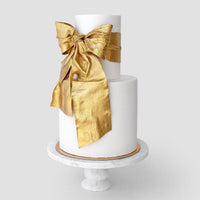 A two-tier white cake on a marble stand features a large bow on the top tier, shimmering with Jenna Rae Cakes Antique Gold Edible Lustre Dust (20g, pack of 3).
