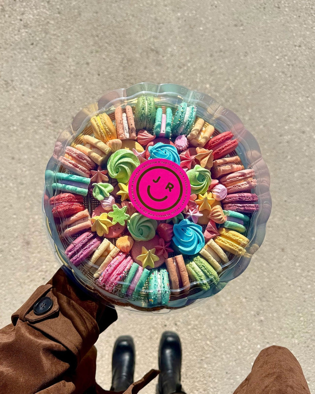 A person in a brown coat and black boots holds a Jenna Rae Cakes Assorted Party Platter—round box of colorful macarons and meringues with a clear lid featuring a pink sticker with a smiling face logo, viewed from above.