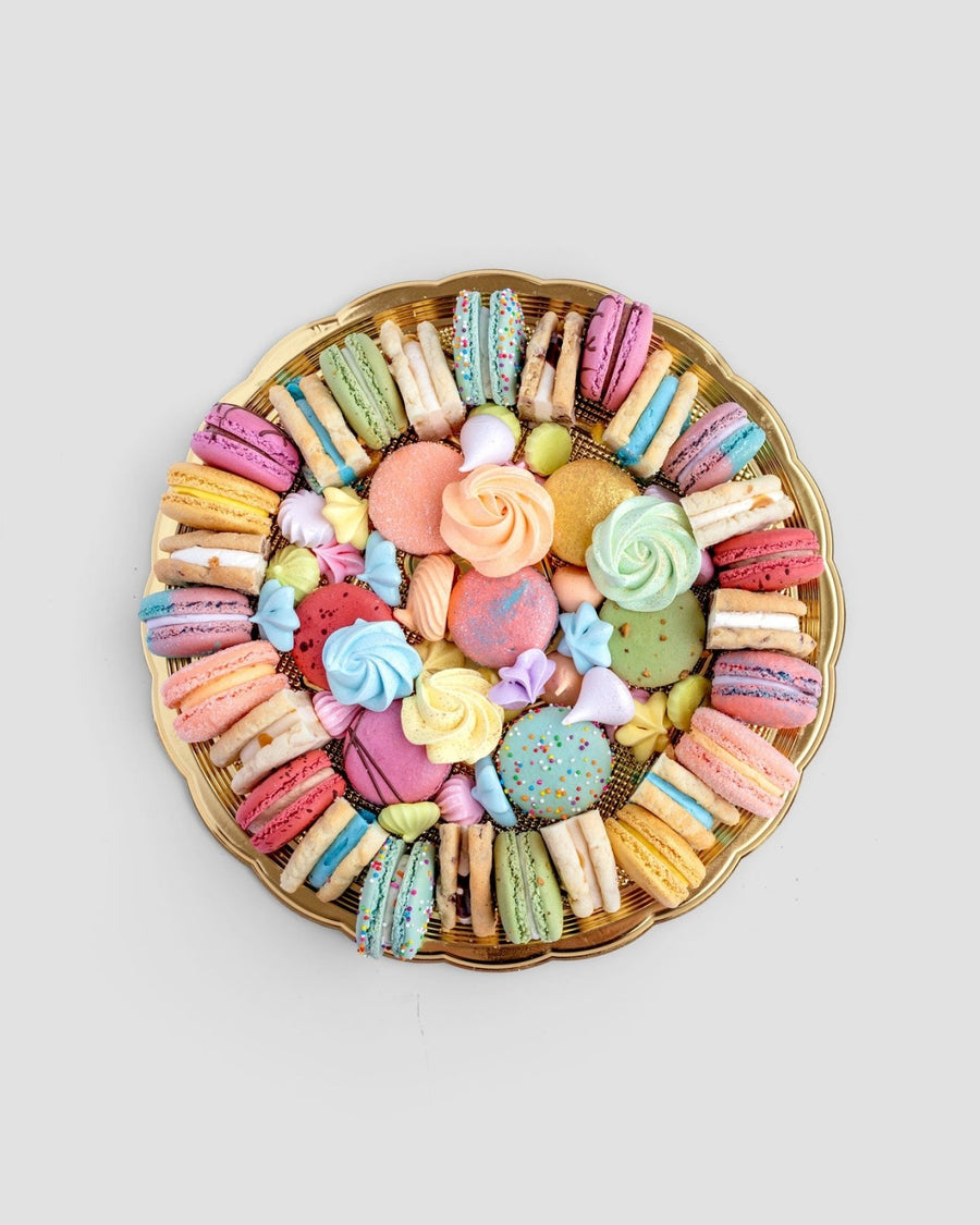 The Jenna Rae Cakes Assorted Party Platter features a round gold tray with colorful macarons, meringues, cookies, and biscuits artfully arranged in a circle on a light gray background.