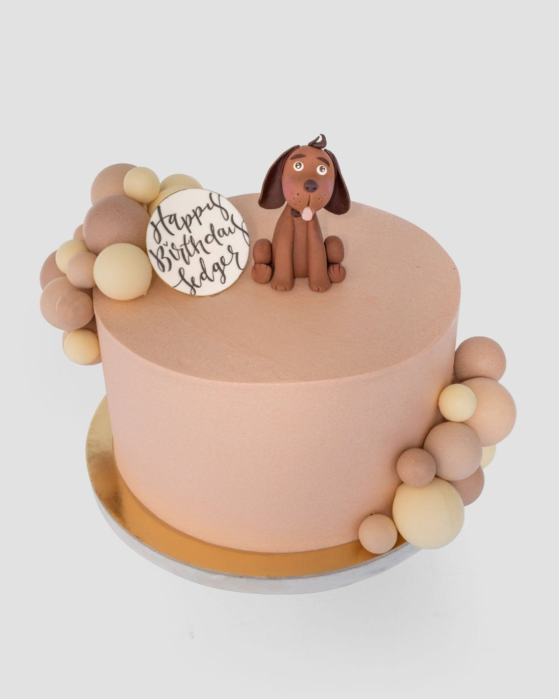 The Baby Animal Balloon Cake by Jenna Rae Cakes is a round, peach-colored birthday cake adorned with brown and beige fondant balls, a fondant dog topper, and a white plaque that says Happy Birthday Edgar.