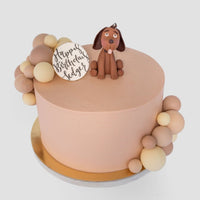 The Baby Animal Balloon Cake by Jenna Rae Cakes is a round, peach-colored birthday cake adorned with brown and beige fondant balls, a fondant dog topper, and a white plaque that says Happy Birthday Edgar.