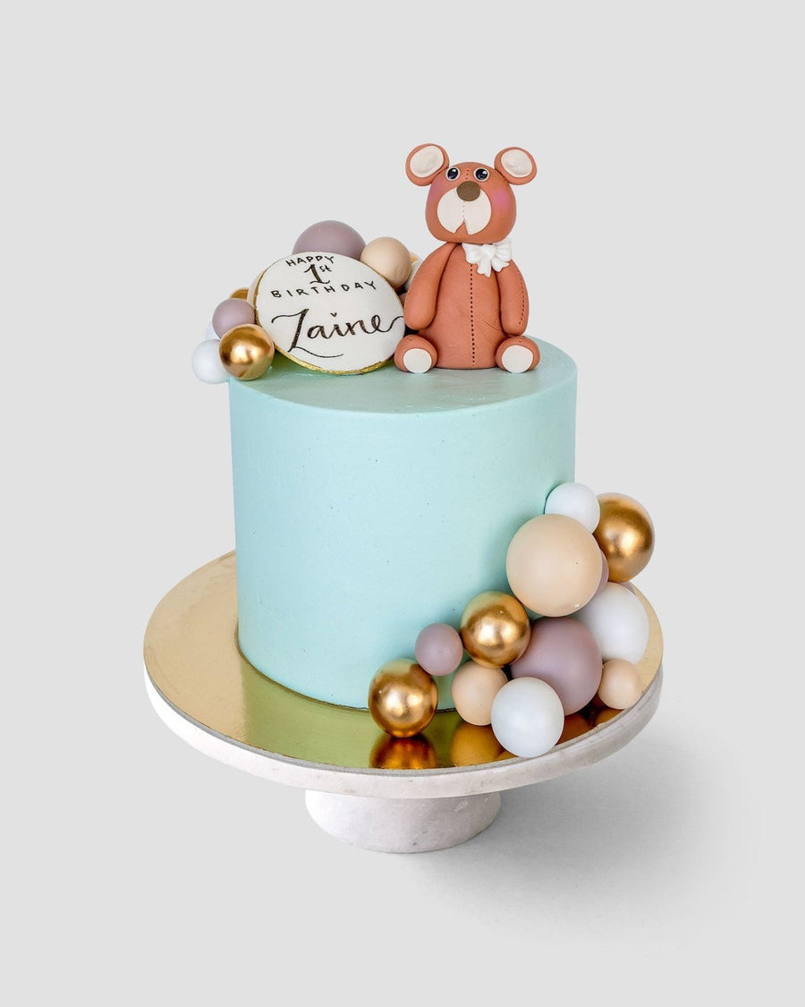 The Baby Animal Balloon Cake by Jenna Rae Cakes features light blue icing, a fondant bear topper, a white plaque with Happy 1st Birthday Taine, and pastel and gold spheres, all displayed on a cake stand.