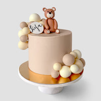 The Baby Animal Balloon Cake by Jenna Rae Cakes is a round beige cake with brown and cream fondant balls, a fondant teddy bear topper, and a “tiya” plaque. It sits on a gold board and white cake stand.