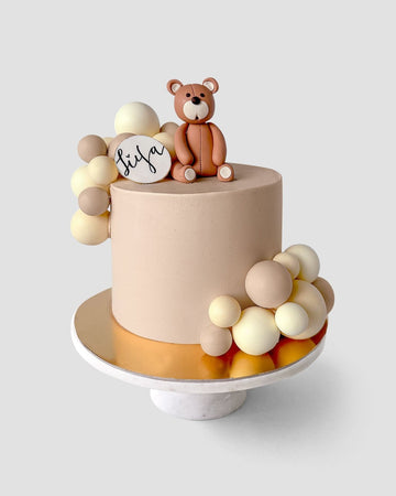 The Baby Animal Balloon Cake by Jenna Rae Cakes is a round beige cake with brown and cream fondant balls, a fondant teddy bear topper, and a “tiya” plaque. It sits on a gold board and white cake stand.