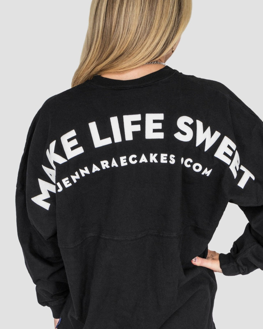 A person with long blonde hair, seen from behind, wears the Jenna Rae Cakes Black + White Spirit Jersey featuring MAKE LIFE SWEET and jennaraecakes.com in bold white letters on the back.