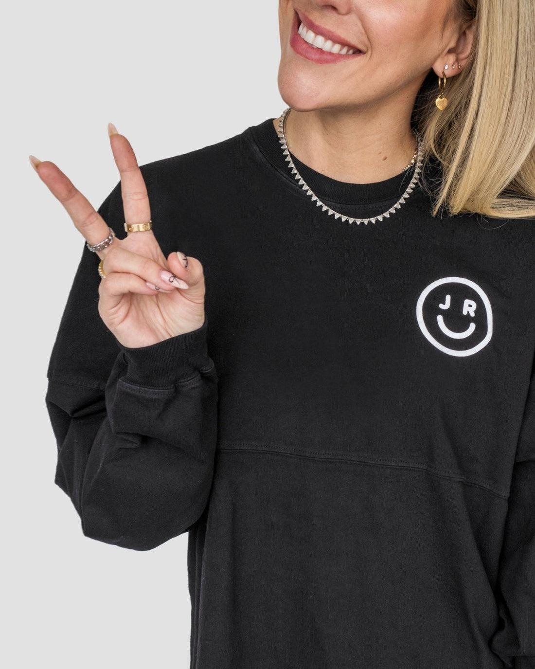 A woman wearing the Black + White Spirit Jersey by Jenna Rae Cakes, featuring a white smiley face and J R logo, flashes a peace sign. She accessorizes with gold rings, hoop earrings, and a chain necklace. Only her upper body is visible.