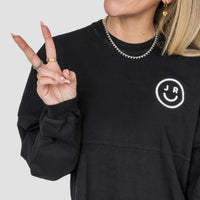 A woman wearing the Black + White Spirit Jersey by Jenna Rae Cakes, featuring a white smiley face and J R logo, flashes a peace sign. She accessorizes with gold rings, hoop earrings, and a chain necklace. Only her upper body is visible.