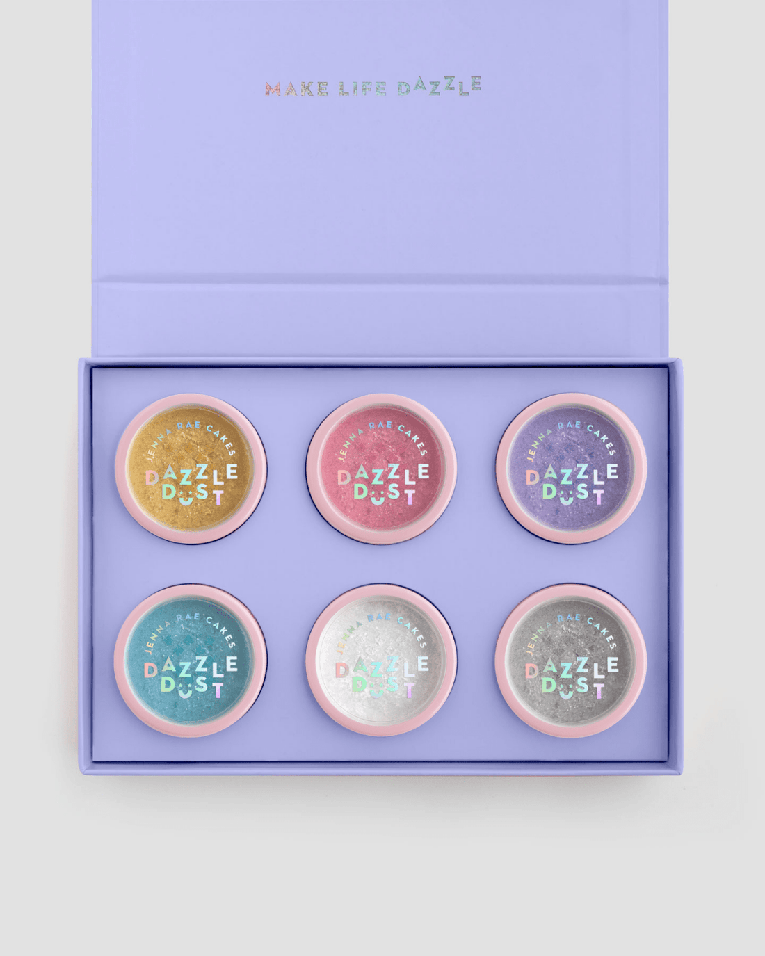 The Build Your Own Dazzle Dust Collection by Jenna Rae Cakes features six round pastel containers labeled Dazzle Dust, arranged in two rows of three inside a lavender gift box with MAKE LIFE DAZZLE on the lid.