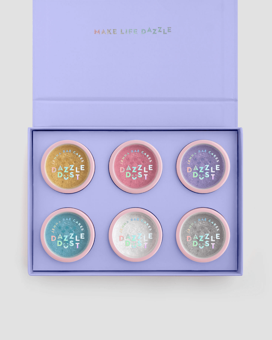 The Build Your Own Dazzle Dust Collection by Jenna Rae Cakes features six round pastel containers labeled Dazzle Dust, arranged in two rows of three inside a lavender gift box with MAKE LIFE DAZZLE on the lid.