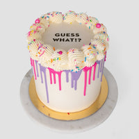 The Burn Away Cake by Jenna Rae Cakes is a round cake with white frosting, colorful sprinkles, pink and purple icing drips, piped cream on top, and GUESS WHAT!? in black letters. It’s displayed on a gold board and marble platter.