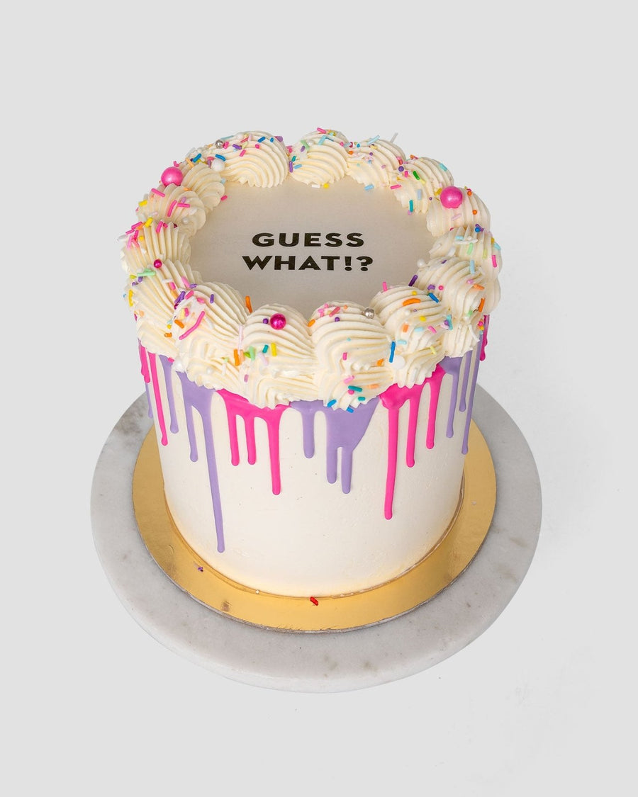 The Burn Away Cake by Jenna Rae Cakes is a round cake with white frosting, colorful sprinkles, pink and purple icing drips, piped cream on top, and GUESS WHAT!? in black letters. It’s displayed on a gold board and marble platter.