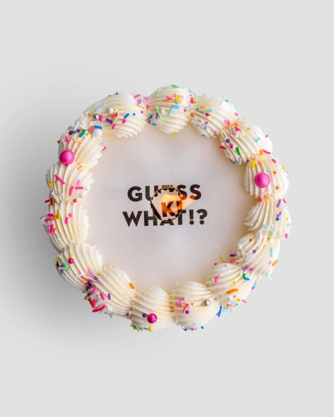 The Burn Away Cake by Jenna Rae Cakes is a round cake with white frosting, colorful sprinkles, pearl decorations, and GUESS WHAT!? written on top with a small orange heart partially covering the middle word.