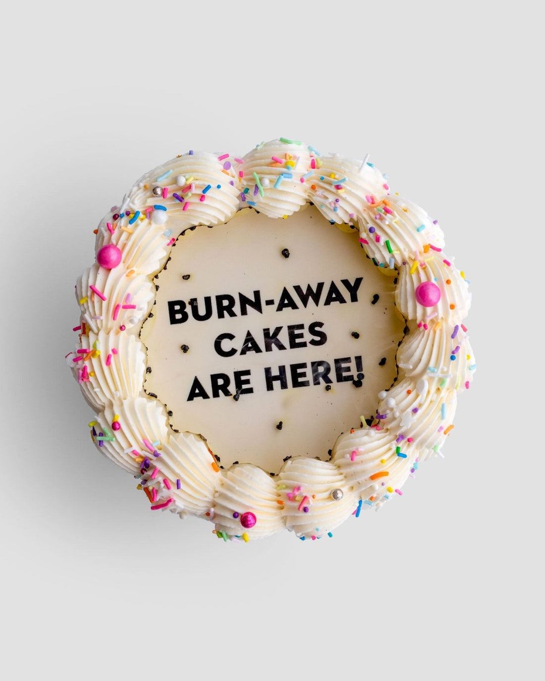 A round Jenna Rae Cakes Burn Away Cake with white frosting, colorful sprinkles, and pink accents. The center displays BURN-AWAY CAKES ARE HERE! in black icing on a smooth white background.
