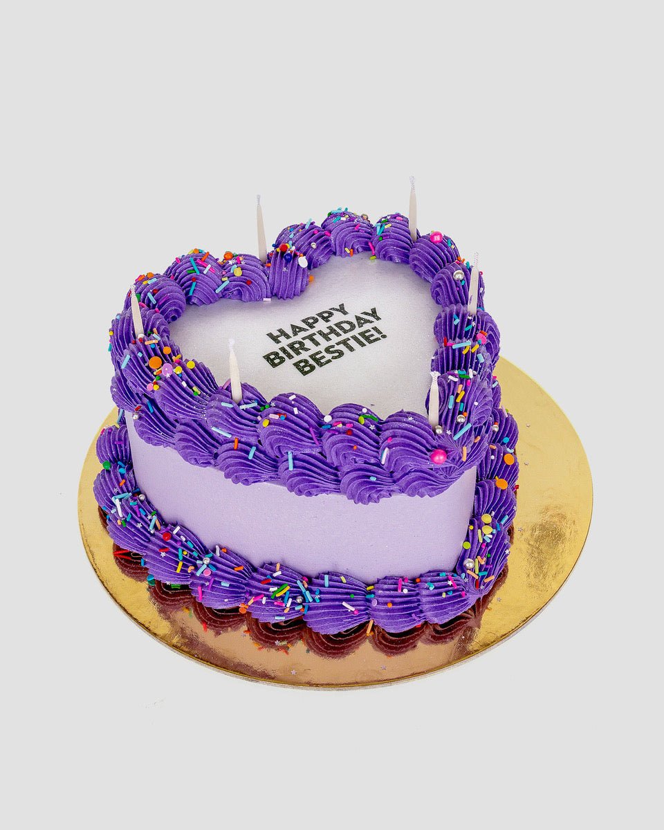 The Burn Away Heart Cake by Jenna Rae Cakes features a heart shape, purple frosting, colorful sprinkles, five unlit candles, and HAPPY BIRTHDAY BESTIE! on top. Its set on a gold cake board against a plain backdrop.