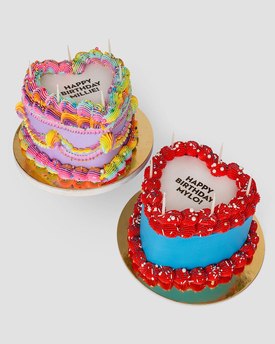 Two Burn Away Heart Cakes by Jenna Rae Cakes: one with rainbow icing and HAPPY BIRTHDAY BITCH, the other with blue and red icing and HAPPY BIRTHDAY MY LOVE, both topped with candles on gold boards.