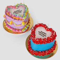 Two Burn Away Heart Cakes by Jenna Rae Cakes: one with rainbow icing and HAPPY BIRTHDAY BITCH, the other with blue and red icing and HAPPY BIRTHDAY MY LOVE, both topped with candles on gold boards.