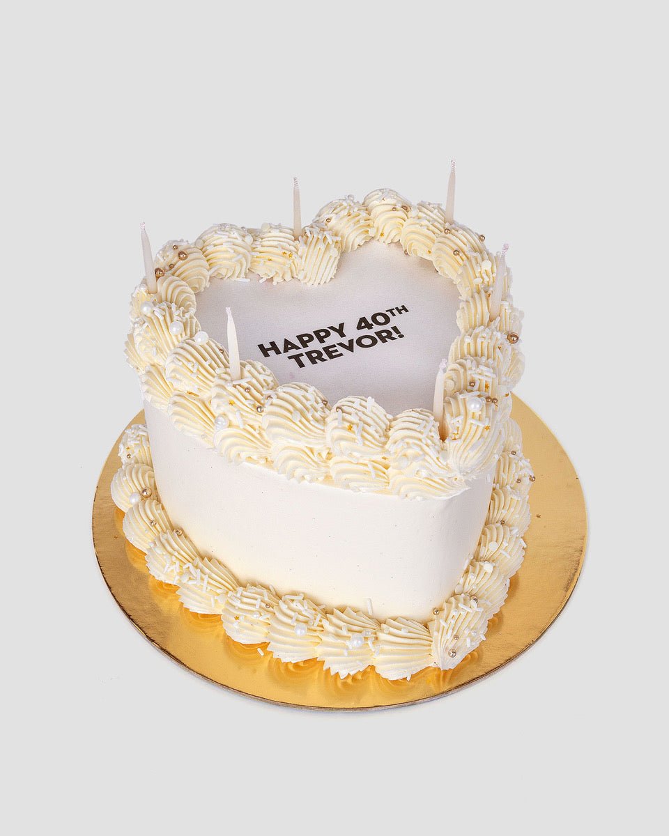 The Burn Away Heart Cake by Jenna Rae Cakes is a heart-shaped white cake with piped cream, pearl-like accents, and five unlit candles. HAPPY 40TH TREVOR! is written on top, displayed on a round gold board against a plain background.