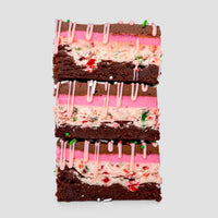 Three stacked Jenna Rae Cakes Candy Cane Brownie bars—limited edition—feature chocolate layers, pink frosting, white cream with red and green sprinkles, and a pink icing drizzle, set against a plain light background.