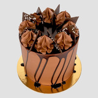 The Chocolate Lovers Cake by Jenna Rae Cakes is a tall, round cake with smooth frosting, chocolate drip, chocolate cream swirls, chocolate shards, and white sprinkles on a gold board against a plain background.