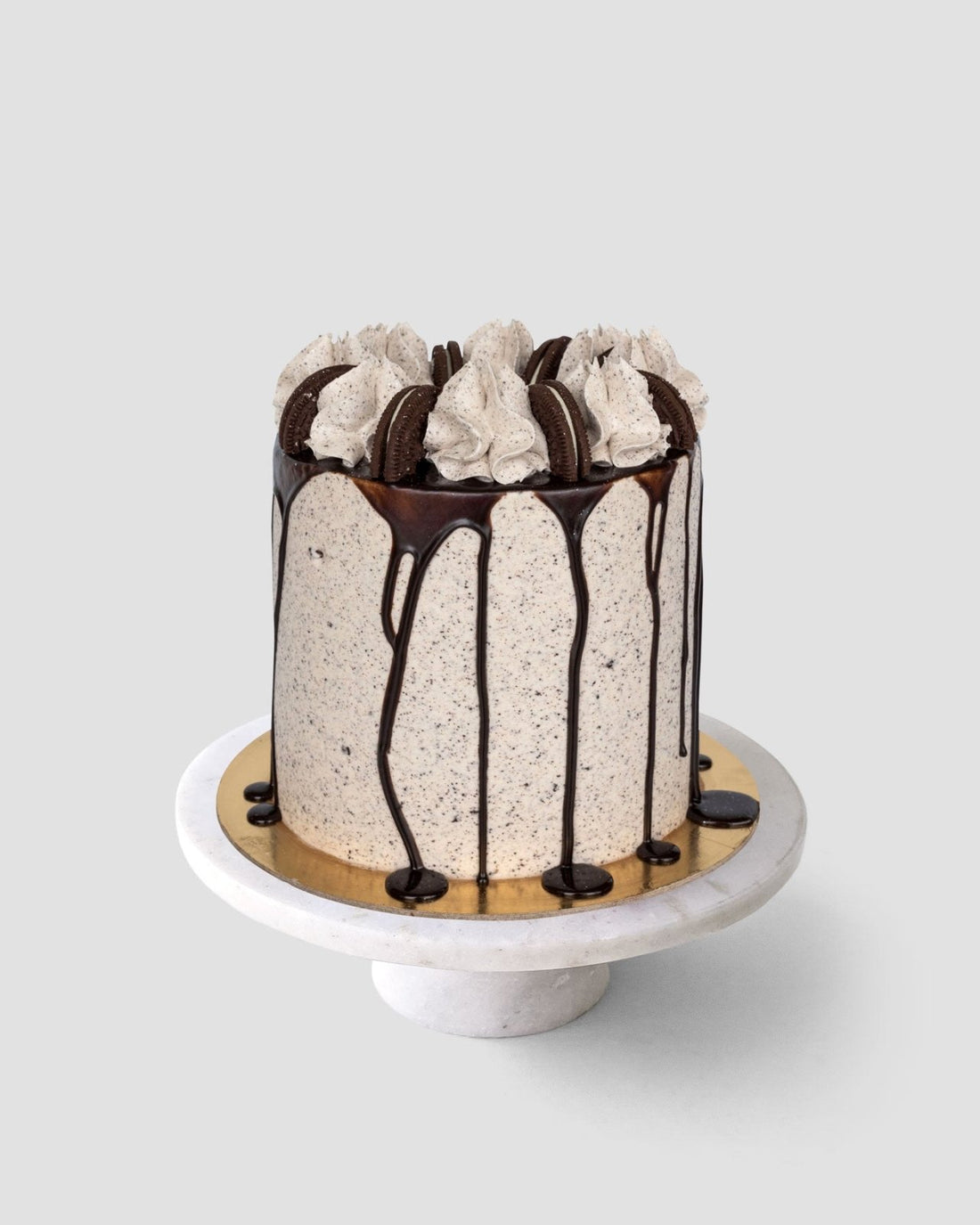 The Chocolate Oreo Cake by Jenna Rae Cakes is a tall cake topped with swirls of cream, chocolate cookies, and chocolate ganache drips, displayed on a white marble cake stand against a plain gray background.