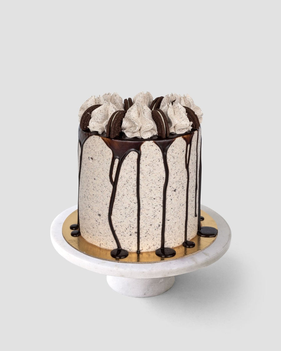The Chocolate Oreo Cake by Jenna Rae Cakes is a tall cake topped with swirls of cream, chocolate cookies, and chocolate ganache drips, displayed on a white marble cake stand against a plain gray background.
