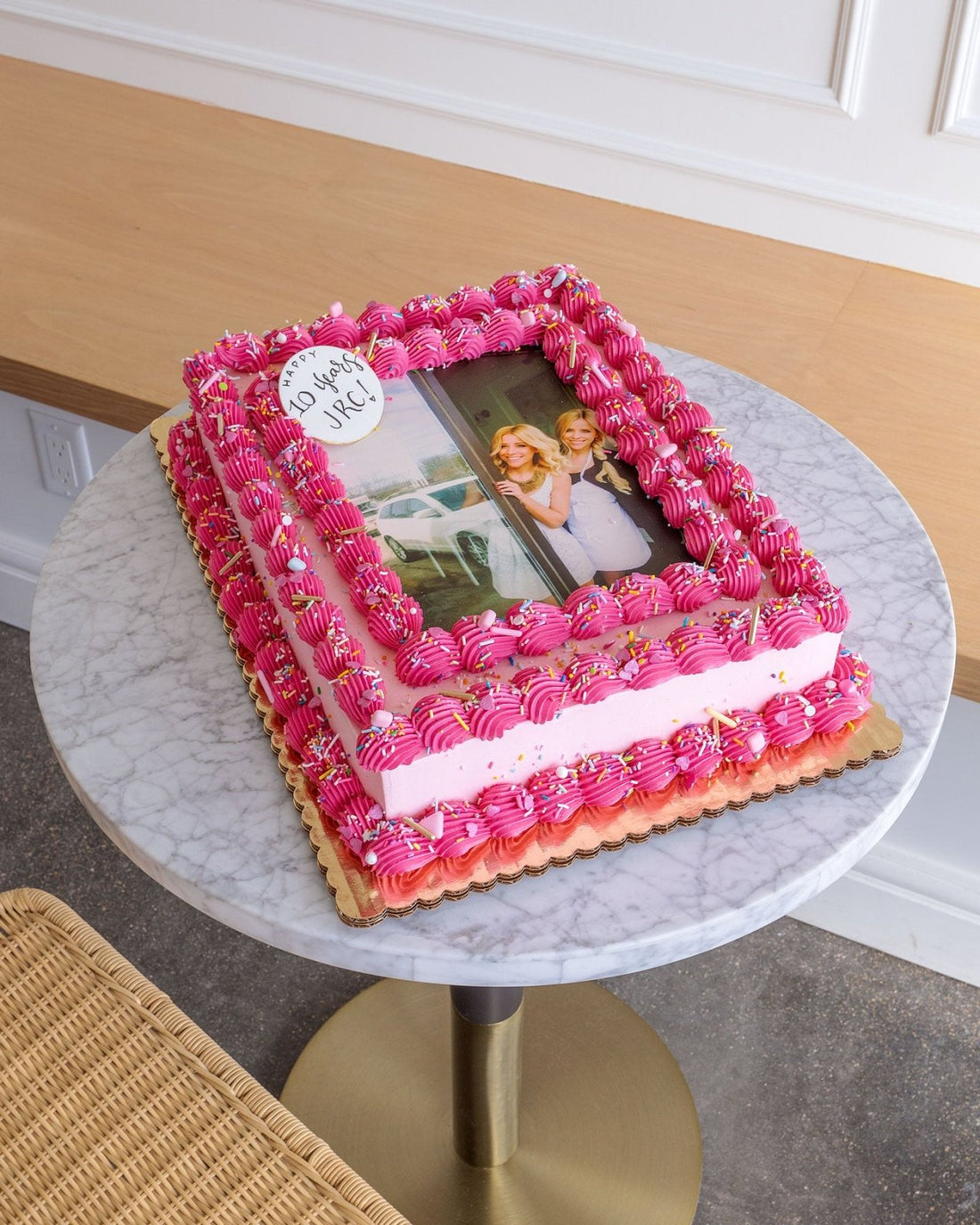 A Classic Sheet Cake by Jenna Rae Cakes with pink frosting and piped borders displays a printed photo of two smiling women. A white plaque on top reads 50 years, NRC! The cake rests on a marble table.